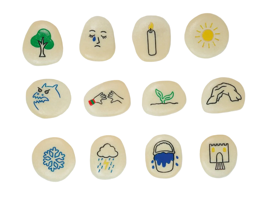 A set of twelve smooth, polished river stones arranged in a 3x4 grid on a white background. Each stone is hand-painted with a simple illustration representing a season or concept: a green tree, a sad face with a tear, a burning candle, a yellow sun, a shark, hands washing, a green sprout, a stone arch, a blue snowflake, rain with lightning, a blue bucket, and a stone castle.