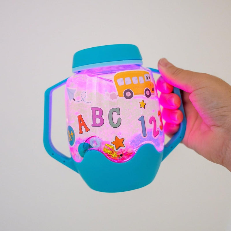 Hand holding a blue Glo Pals sensory jar glowing pink, decorated with ABC letters, numbers and a school bus