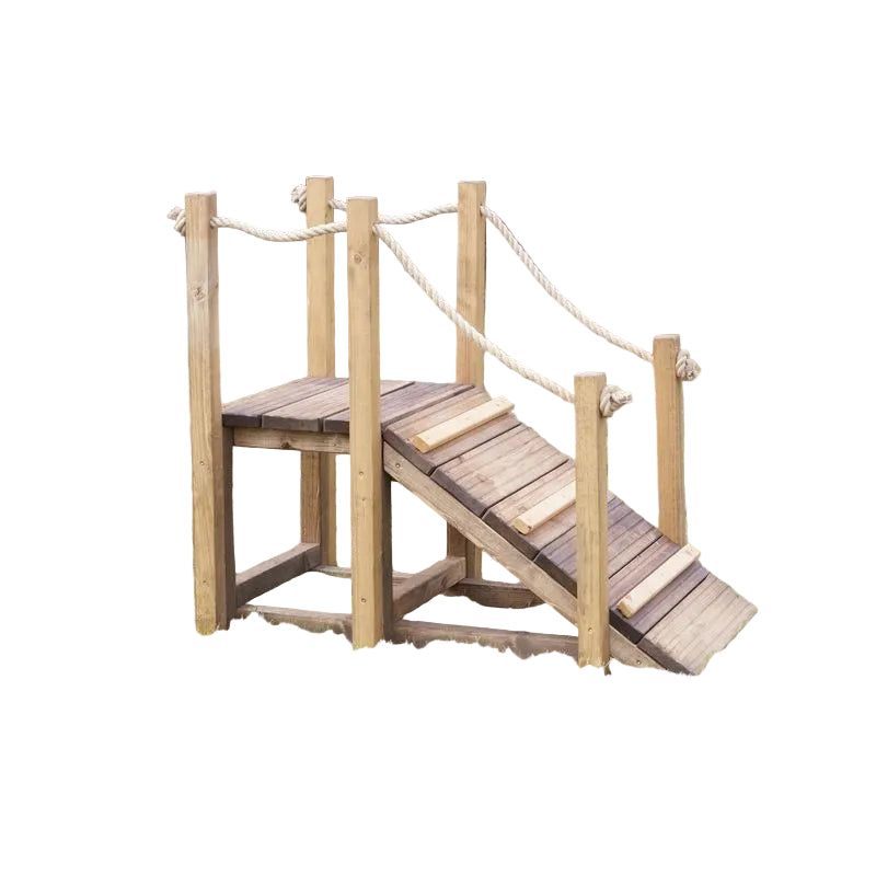 A wooden outdoor play bridge with a ramp and rope railings, set on a grassy lawn with trees in the background.