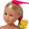 A blonde doll with blue eyes shown in a close-up, with a bright pink hairbrush resting on her hair and a pink hair tie at the back of her head. A decorative badge in the lower right says 'BRUSH AND STYLE'.