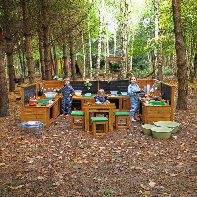Three children in blue patterned clothes play with a large wooden outdoor cooking and play kitchen set in a wooded campsite setting.