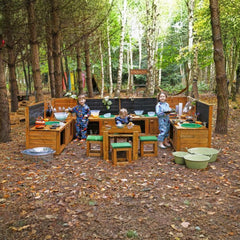 Three children in blue patterned clothes play with a large wooden outdoor cooking and play kitchen set in a wooded campsite setting.