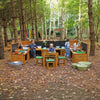 Three children in patterned overalls play at an outdoor wooden play kitchen set in a wooded forest clearing with leaf-covered ground.