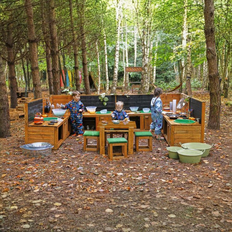 Three children in patterned overalls play at an outdoor wooden play kitchen set in a wooded forest clearing with leaf-covered ground.