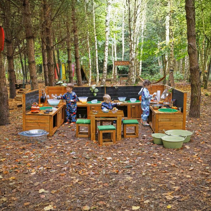 Three children in blue patterned overalls play with a wooden outdoor cooking and play set in a wooded forest area with fallen leaves on the ground.