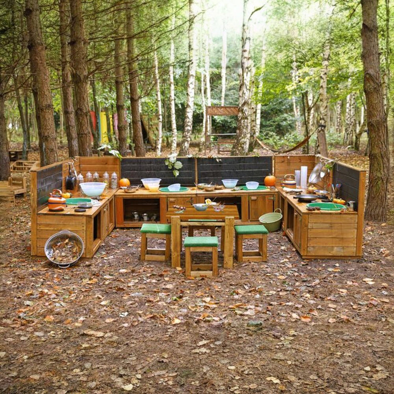 Outdoor wooden camp kitchen set with cooking stations, bowls, bottles, and a small table with green cushioned stools, arranged in a forest clearing with trees and leaf-covered ground.