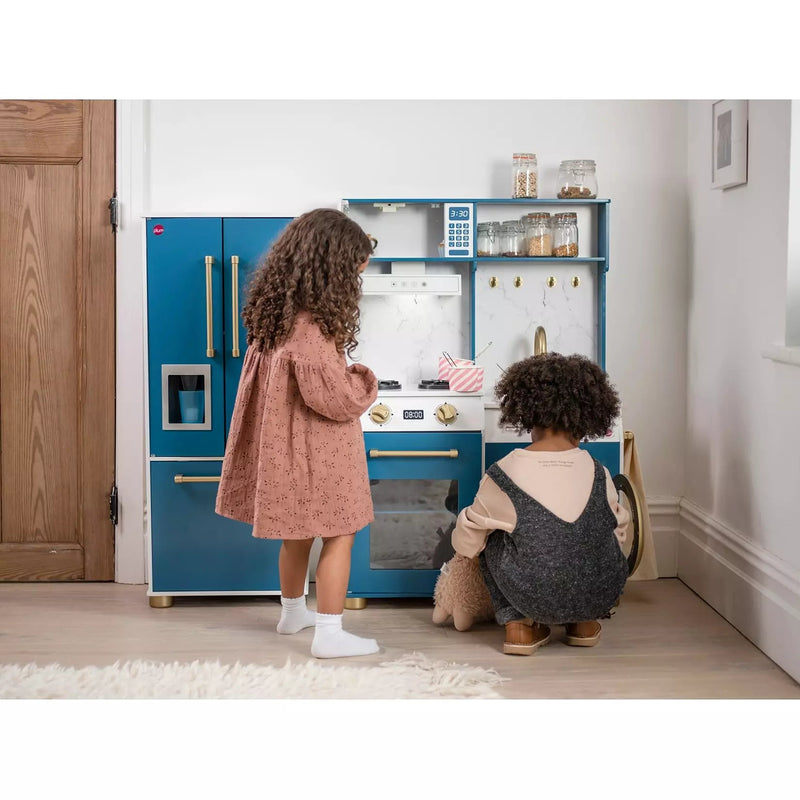 Two children playing with a blue toy kitchen set in a bright room. One child stands in a dusty pink dress, and the other crouches in dark gray overalls next to a teddy bear.